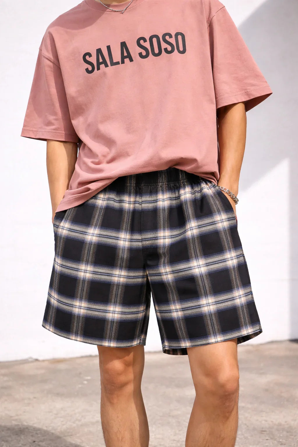 Man wearing a pink 'SALA SOSO' t-shirt and black plaid shorts outdoors