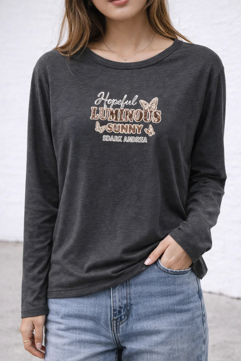 Woman wearing dark gray long sleeve graphic t-shirt with butterfly and hopeful luminous sunny text, paired with blue jeans.